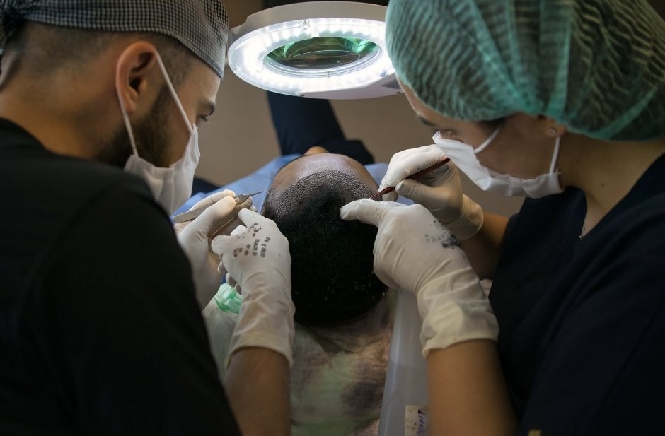 Two clinicians in scrubs and masks working together under a magnifying lamp to place grafts into a patient's scalp.